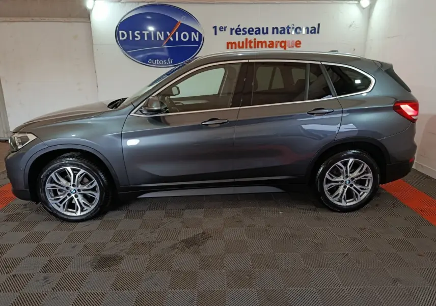 BMW X1 sDrive18i Lounge gris foncé vue de profil côté gauche dans un garage avec logo Distinxion en arrière-plan.