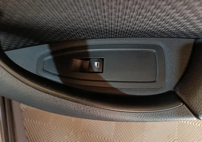 Gros plan sur le bouton de commande de lève-vitre noir dans l'accoudoir intérieur d'une BMW X1 gris foncé.