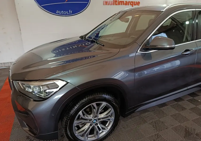 BMW X1 sDrive18i gris foncé vue 3/4 avant côté gauche, avec jante alliage et phares LED distinctifs.