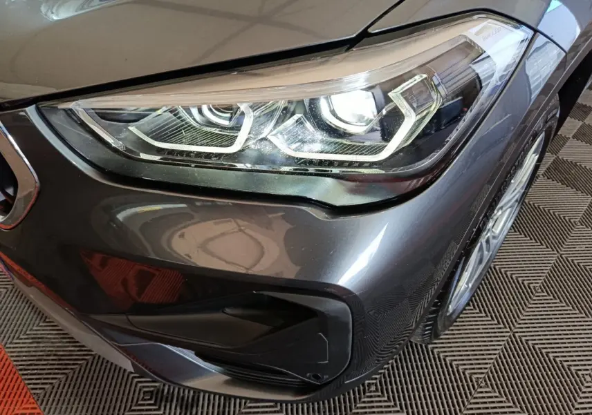 Gros plan sur le phare avant droit d’un BMW X1 sDrive18i Lounge gris foncé avec jante et partie du pare-chocs visibles.