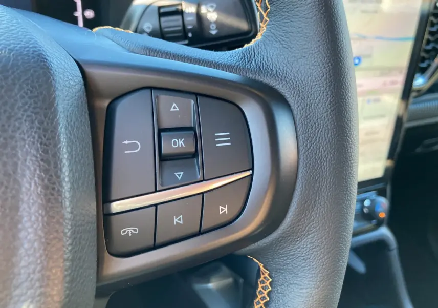 Gros plan sur les commandes multifonctions à gauche du volant cuir noir du Ford Ranger Wildtrak blanc glacier, avec écran tactile en arrière-plan.
