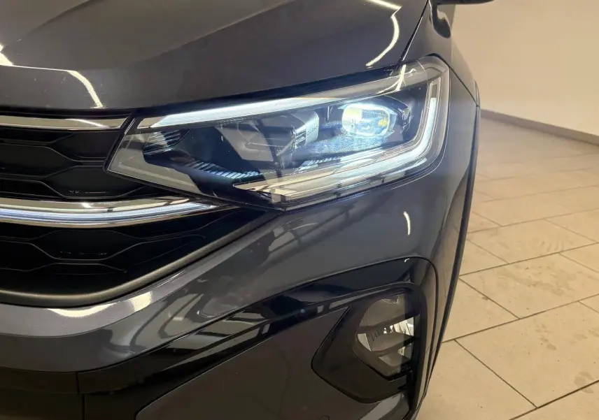 Gros plan sur le phare avant droit du Volkswagen Taigo gris cendré 2024 avec détails lumineux et calandre noire brillante.