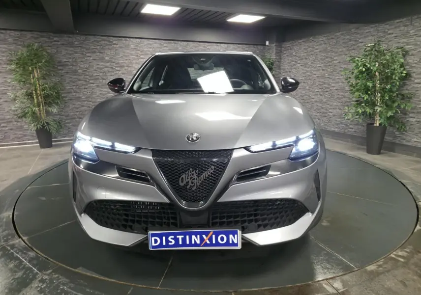 Vue avant d'une Alfa Romeo Junior 1.2 Ibrida gris métallisé avec calandre noire et phares LED allumés.