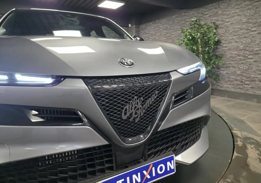 Vue avant rapprochée d'une Alfa Romeo Junior 1.2 Ibrida gris métallisé avec calandre noire et phares LED allumés.