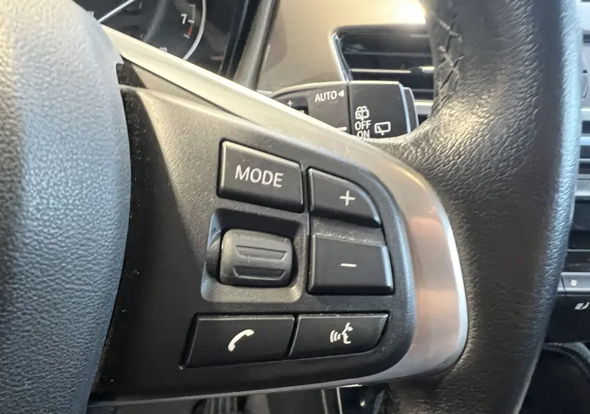 Gros plan sur les commandes au volant gauche du BMW X1 F48 noir, avec boutons mode, volume et téléphone visibles.