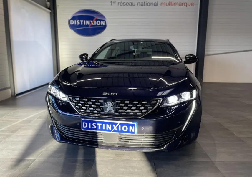 Peugeot 508 SW bleu foncé vue de face en intérieur, avec calandre distinctive et phares allumés.