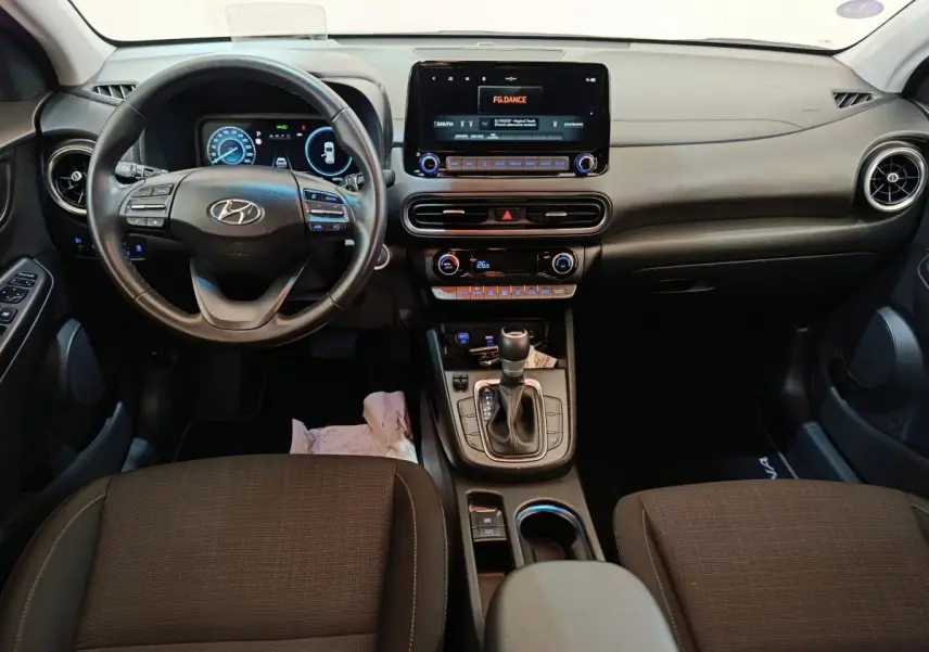 Vue intérieure avant du Hyundai Kona 1.6 Hybrid 2023, tableau de bord noir avec écran tactile central et volant cuir multifonctions.