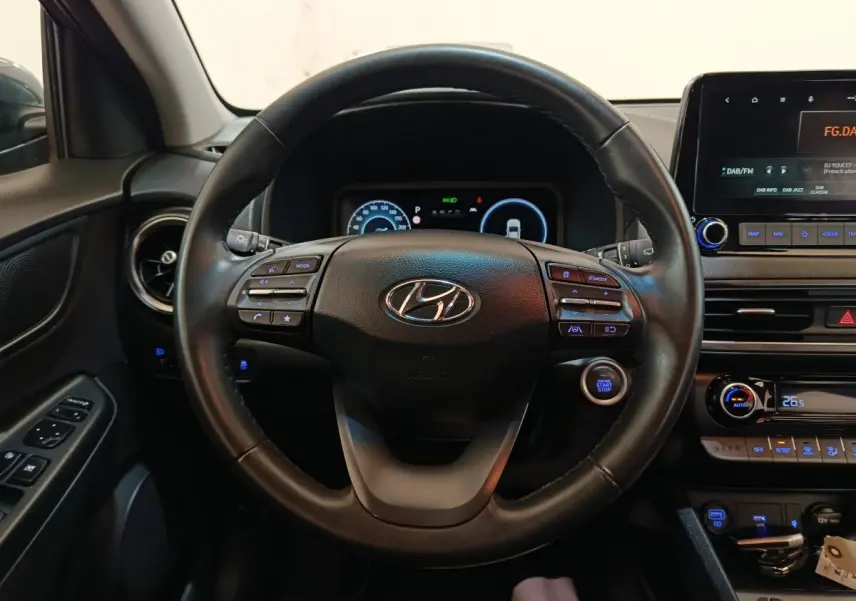 Vue intérieure centrée sur le volant cuir multifonctions d'une Hyundai Kona 1.6 Hybrid 2023 avec tableau de bord numérique.