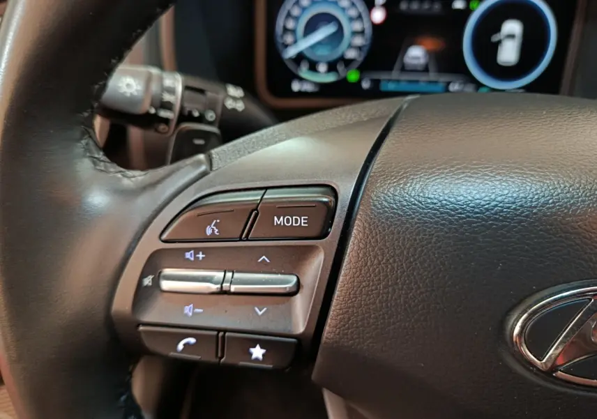 Gros plan sur le volant cuir noir du Hyundai Kona 2023, avec commandes multifonctions et tableau de bord numérique en arrière-plan.