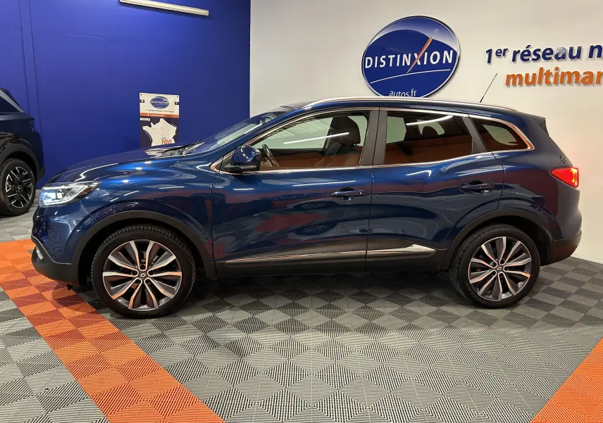 Profil côté gauche d'un Renault Kadjar bleu 2017 avec jantes alliage et vitres teintées en intérieur showroom.