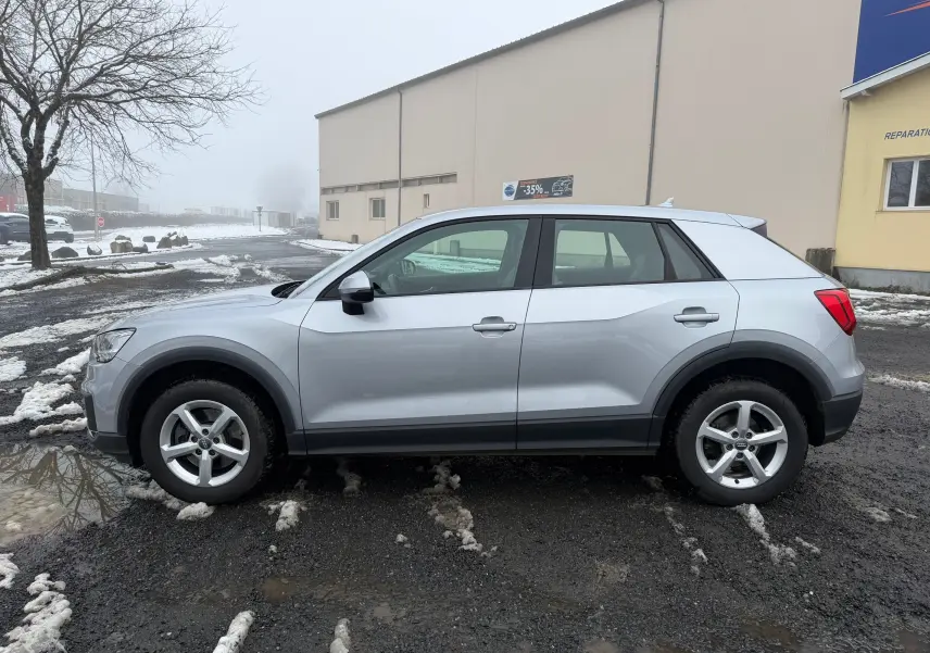 Audi Q2 gris aluminium vue 3/4 avant droit sur parking avec bâtiment AutoCantal en arrière-plan.