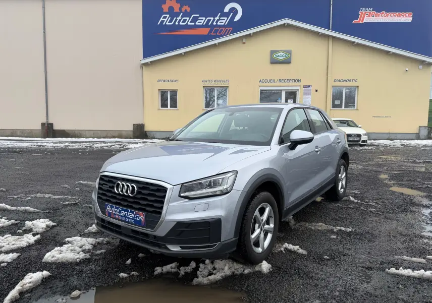 Audi Q2 gris aluminium vue de profil côté gauche sur un sol humide avec traces de neige et bâtiment en arrière-plan.