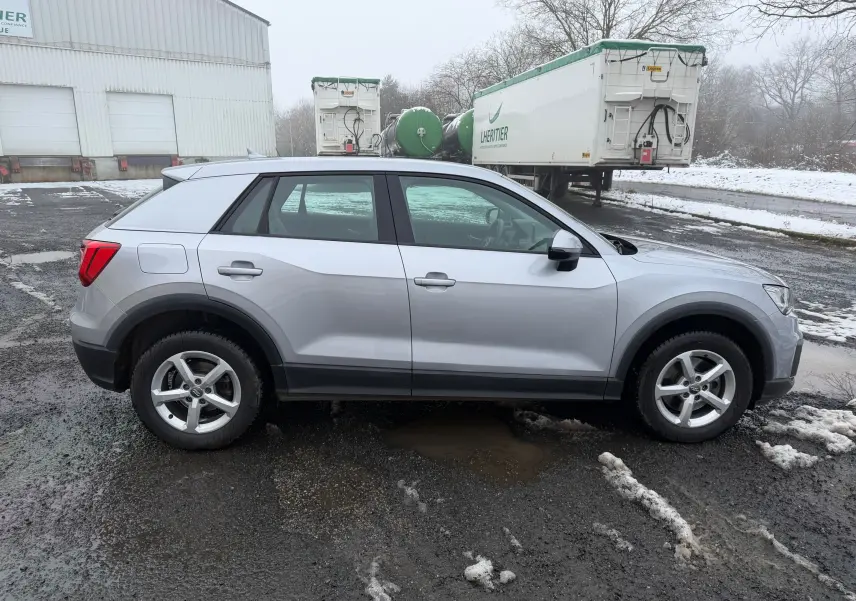 Audi Q2 gris aluminium vu de profil côté gauche sur un sol humide avec arrière-train légèrement surélevé.