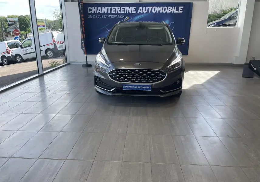 Vue frontale d'un Ford S-MAX Vignale gris magnetic 2022, avec calandre hexagonale et phares LED allumés en showroom.