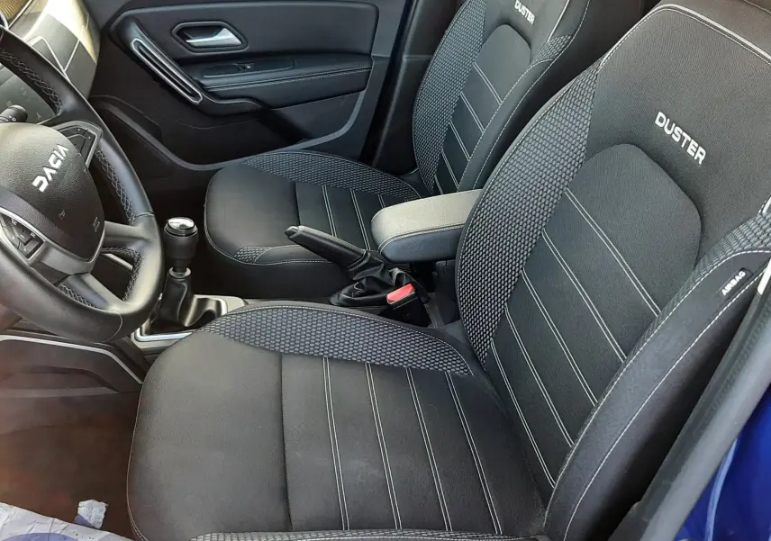 Vue intérieure côté conducteur du Dacia Duster bleu 2023, sièges noirs avec logo DUSTER brodé et volant multifonction.