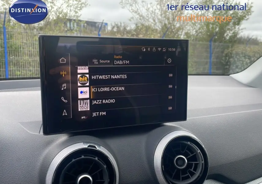 Écran tactile central affichant les stations radio, avec aérateurs ronds argentés sur tableau de bord noir.