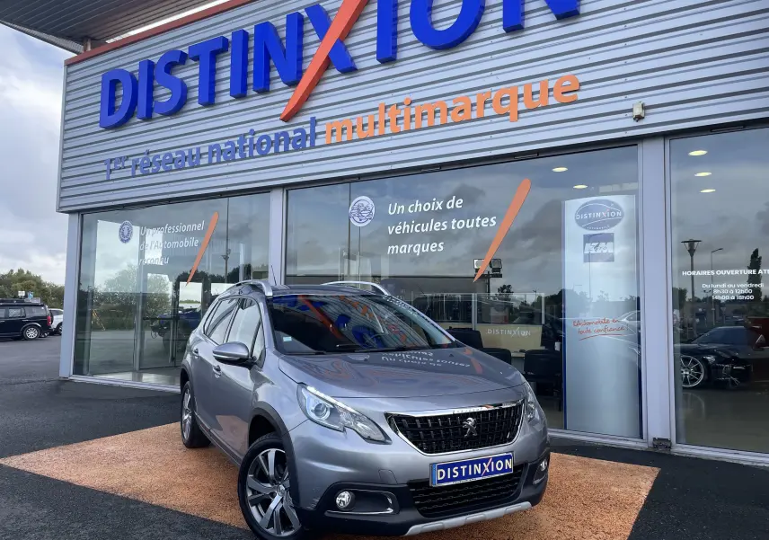 Peugeot 2008 gris en 3/4 avant droit devant une concession Distinxion, avec jantes alliage et calandre noire.