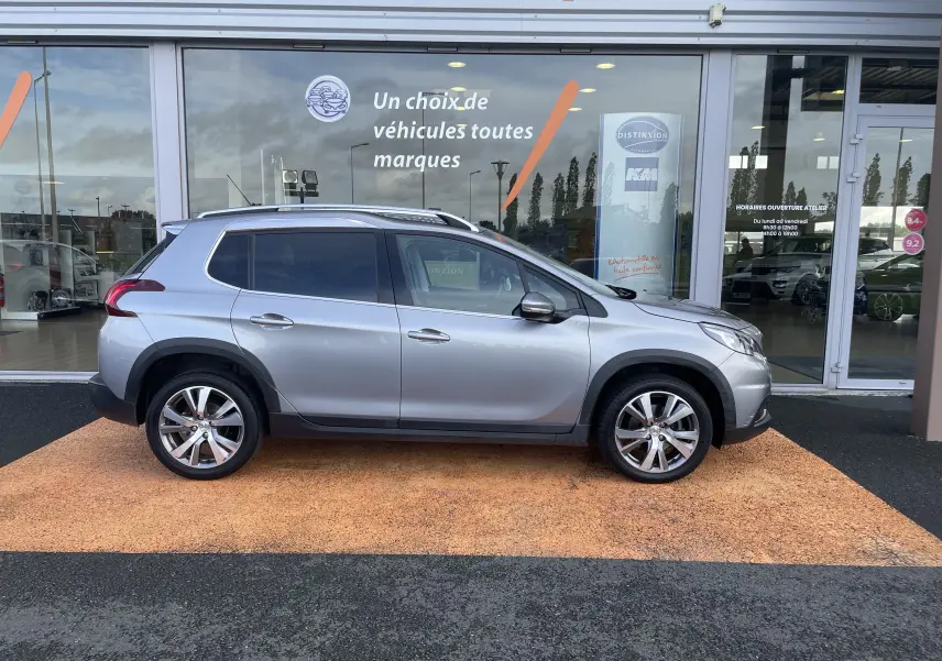 Profil droit d'un Peugeot 2008 gris 2019 garé devant une vitrine de concession avec jantes alliage visibles.