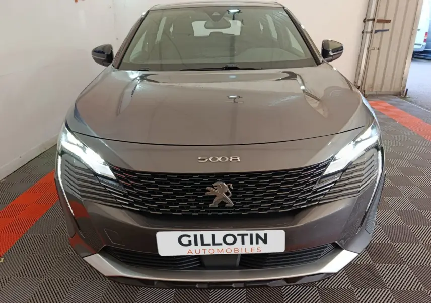 Vue avant d'un Peugeot 5008 gris clair 2023 avec calandre noire et phares LED allumés dans un garage.