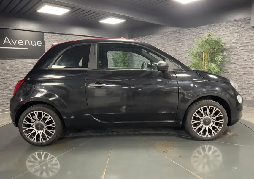 Profil côté gauche de la Fiat 500 noire avec jantes alliage et capote rouge visible en intérieur showroom.