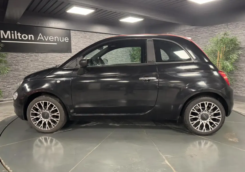 Profil côté gauche d'une Fiat 500 noire avec capote rouge et jantes alliage 16 pouces en intérieur showroom.