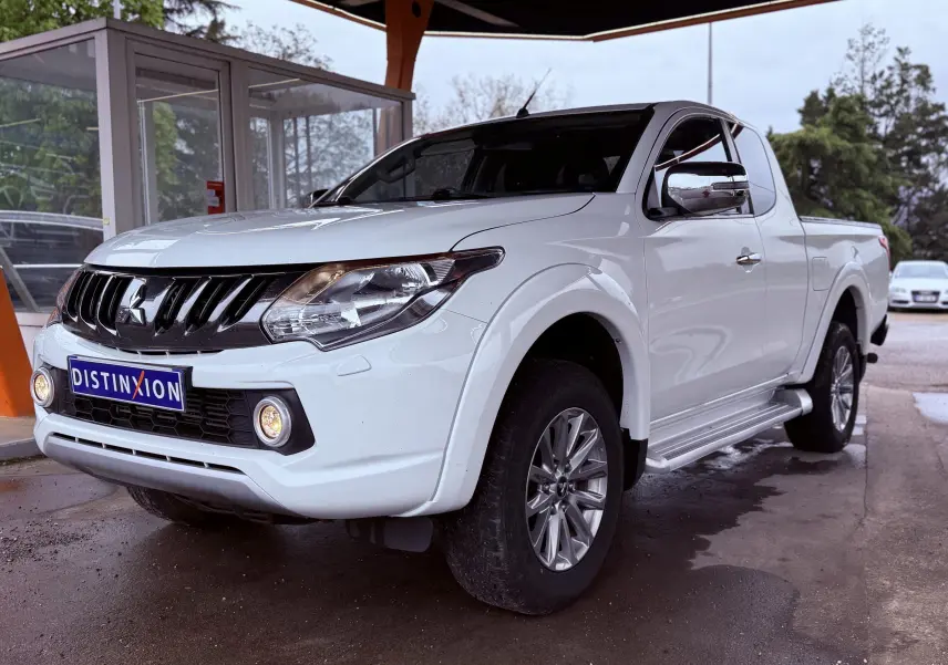 Mitsubishi L200 Club Cab blanc vu en 3/4 avant droit avec calandre noire et phares allumés sous abri.