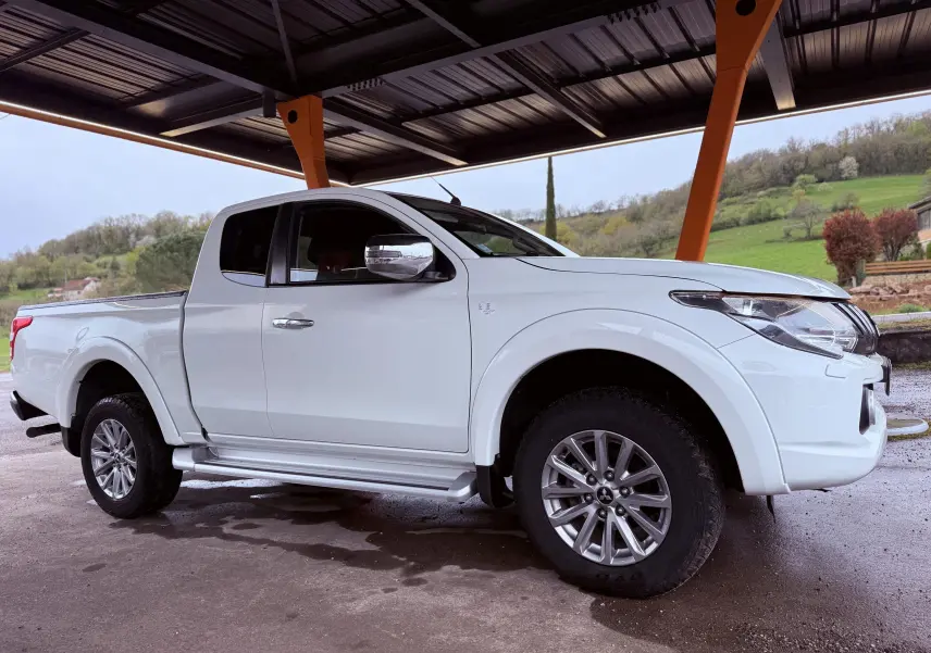 Vue 3/4 avant droit du pick-up Mitsubishi L200 Club Cab blanc sous un abri, avec jantes alliage et marchepieds visibles.