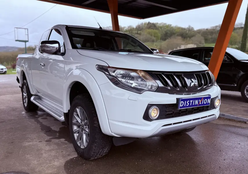 Vue 3/4 avant gauche d’un Mitsubishi L200 Club Cab blanc, avec calandre noire et antibrouillards allumés sous abri.