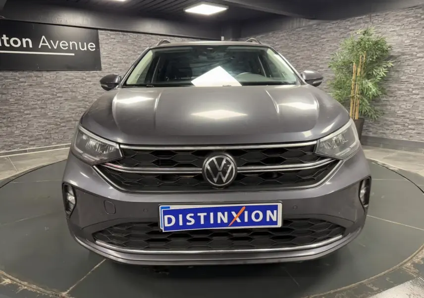 Vue frontale d'un Volkswagen Taigo gris Ascot avec calandre noire et logo VW centré, en showroom intérieur.