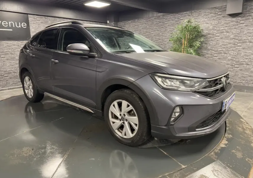 Volkswagen Taigo gris Ascot en 3/4 avant droit, jantes alliage Belmopan et feux LED allumés en intérieur showroom.