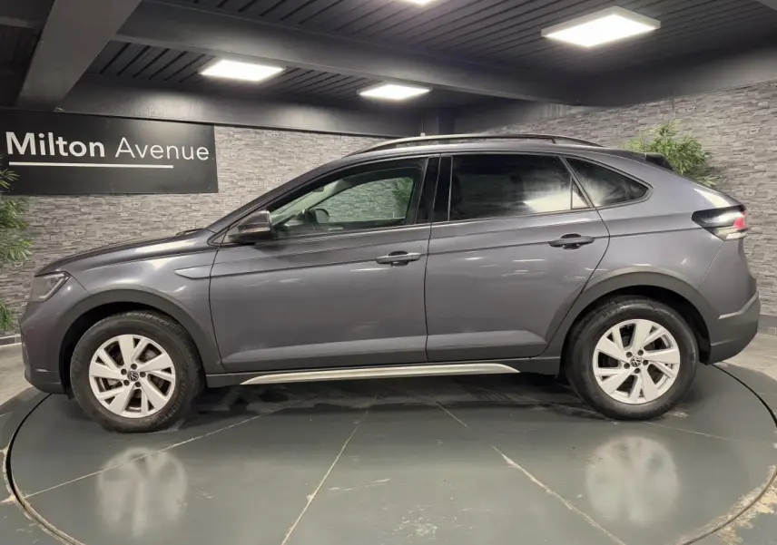 Profil droit d'un Volkswagen Taigo gris 2024 avec jantes alliage Belmopan et barres de toit noires dans un showroom.