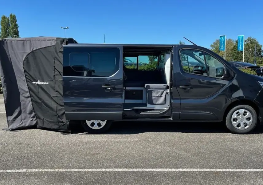 Profil côté droit du Renault Trafic Fourgon gris Comète 2022 avec porte coulissante ouverte et tente d’auvent déployée à l’arrière.