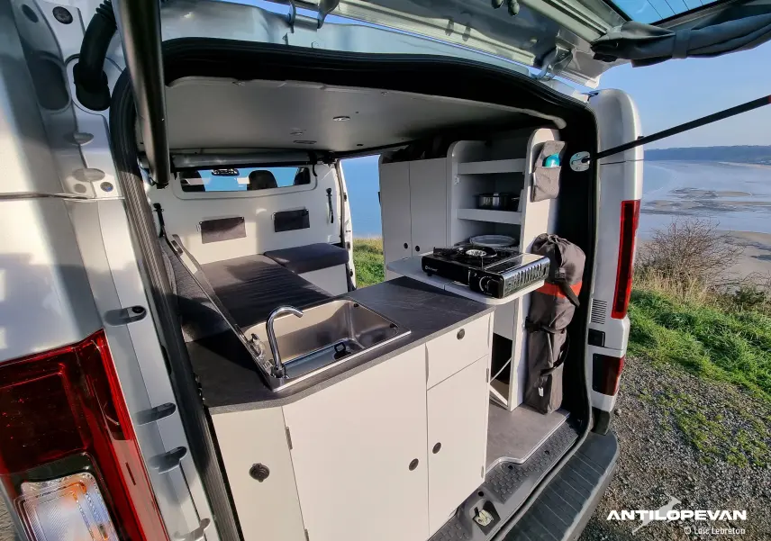 Intérieur arrière du Renault Trafic Fourgon gris comète, montrant cuisine équipée avec évier et réchaud gaz ouvert sur paysage côtier.
