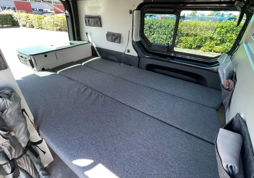 Intérieur du Renault Trafic Fourgon gris Comète, vue latérale droite montrant le lit adulte déplié et rangements muraux.