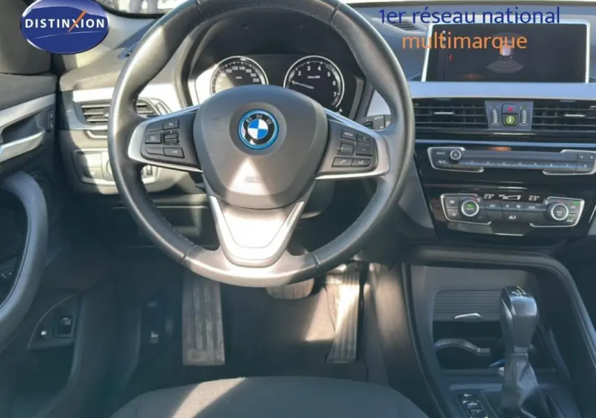Vue intérieure centrée sur le volant sport et la console centrale d'une BMW X1 gris foncé avec écran GPS et boîte auto.