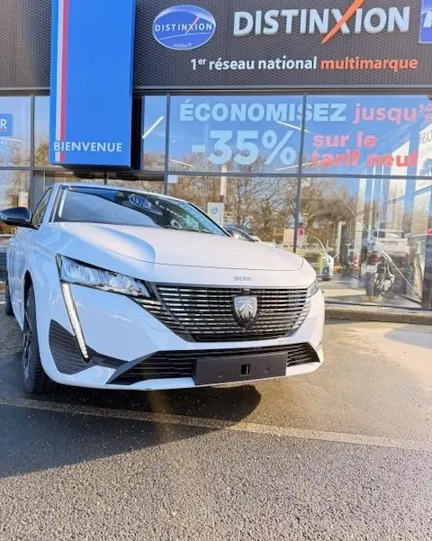 Peugeot 308 Hybrid blanc vue 3/4 avant, calandre noire distinctive et phares LED verticaux, devant concession multimarque.