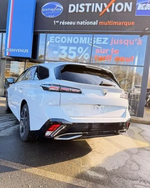Vue 3/4 arrière droite d'une Peugeot 308 Hybrid 145 e-DCS6 Allure blanche avec feux LED et jantes noires devant un concessionnaire multimarque.