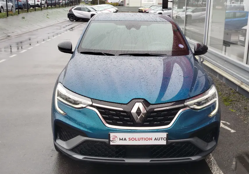 Vue avant d'un Renault Arkana bleu Zanzibar 2022 sous la pluie, avec calandre noire et phares LED allumés.