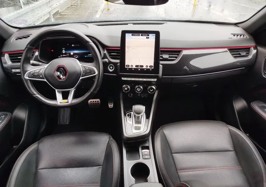 Vue intérieure avant du Renault Arkana 1.6 E-Tech 145ch RS Line 2022, tableau de bord noir avec touches rouges et écran tactile central.