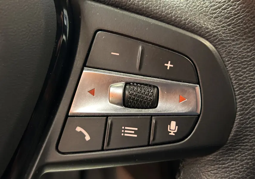 Gros plan sur les commandes au volant côté gauche d'une BMW Série 1 noire 2023, avec molette et boutons multifonctions.