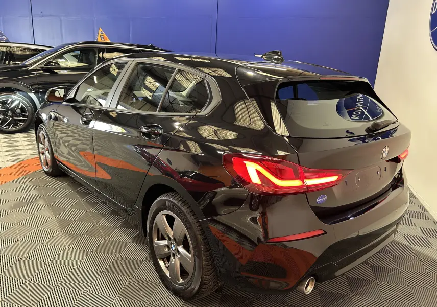 BMW Série 1 16d noire vue 3/4 arrière droit, feux LED allumés et jantes alliage dans un showroom.