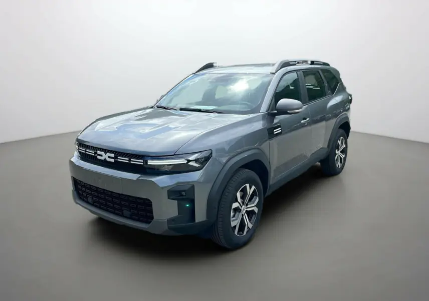 Dacia Bigster gris schiste en 3/4 avant droit, avec barres de toit et jantes alu 17 pouces visibles.