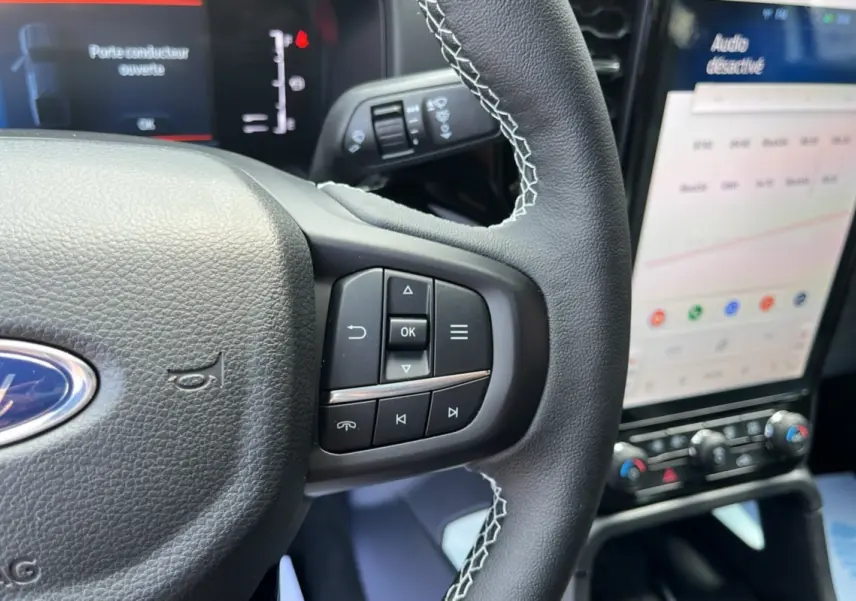 Gros plan sur le volant cuir multifonctions avec coutures blanches du Ford Ranger 2.3 PHEV, écran tactile en arrière-plan.