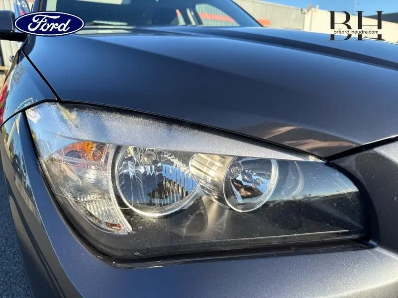 Gros plan sur le phare avant gauche d'une BMW X1 Mineralgrau 2013 avec reflets et détails du bloc optique.