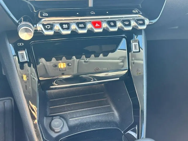 Gros plan sur la console centrale noire brillante de la Peugeot 208 1.2 100ch Allure, avec boutons de commande et prise 12V visible.