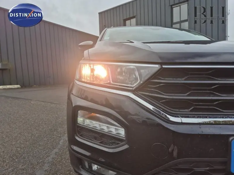 Gros plan sur l'avant gauche gris du Volkswagen T-Roc 1.0 TSI 110ch United avec phare allumé et calandre chromée.