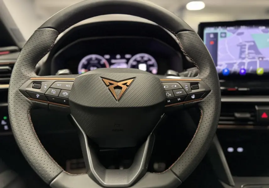 Volant perforé noir avec logo CUPRA cuivre au centre, tableau de bord digital et écran tactile de navigation visible en arrière-plan