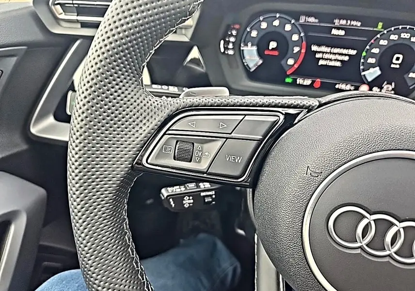 Gros plan sur le volant cuir perforé côté gauche de l'Audi A3 Sportback noir mythos avec tableau de bord digital.