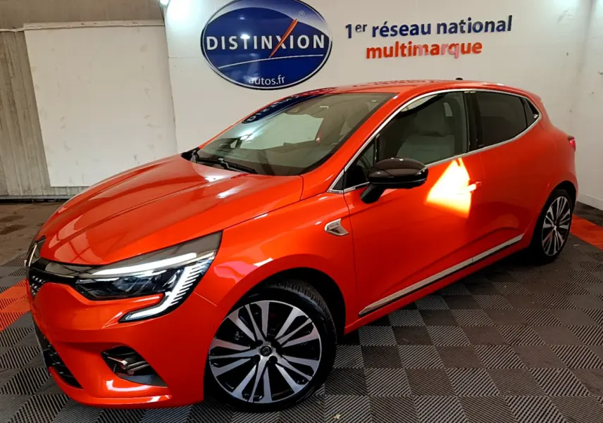 Vue 3/4 avant droite d'une Renault Clio Initiale Paris orange vif dans un showroom avec sol damier noir et rouge.