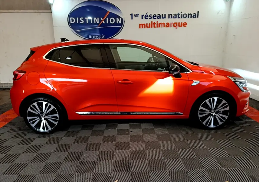 Profil côté droit d'une Renault Clio Initiale Paris orange vif, avec jantes alliage et vitres teintées en intérieur showroom.